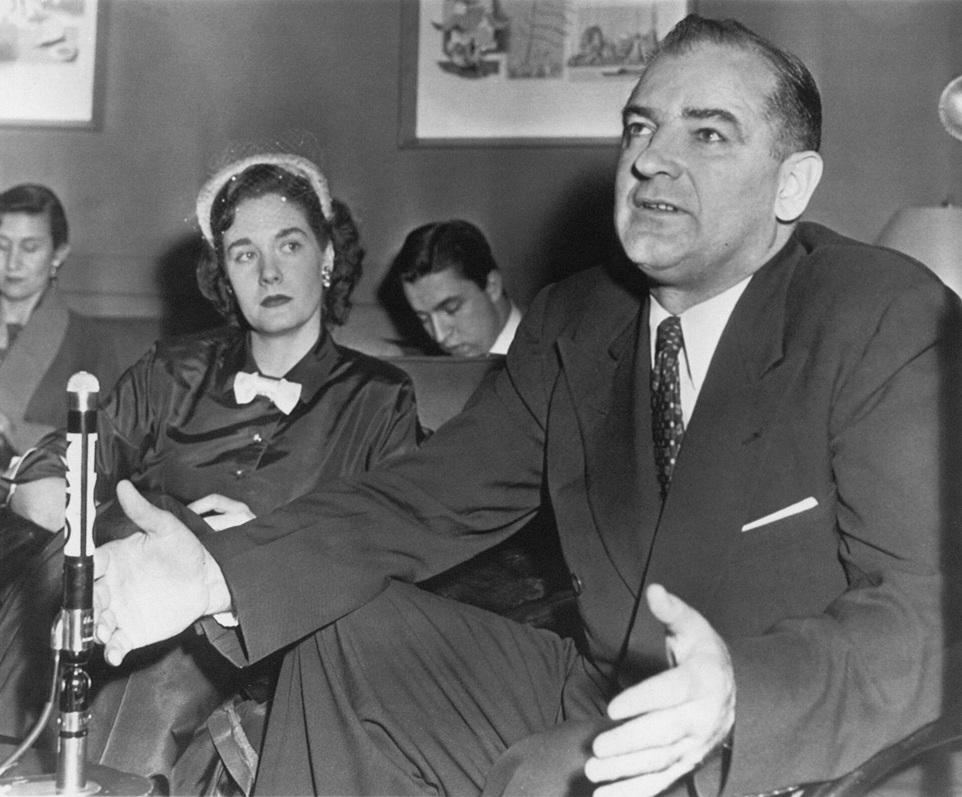 Black and white photograph of Senator Joseph McCarthy seated at a table with a microphone, speaking during a hearing, with a woman seated beside him, February 10, 1954.
